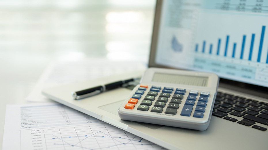 Un ordinateur portable, dont l’écran affiche des données financières, reposant sur un bureau à côté d’une calculatrice, d’un stylo et de rapports statistiques imprimés. 