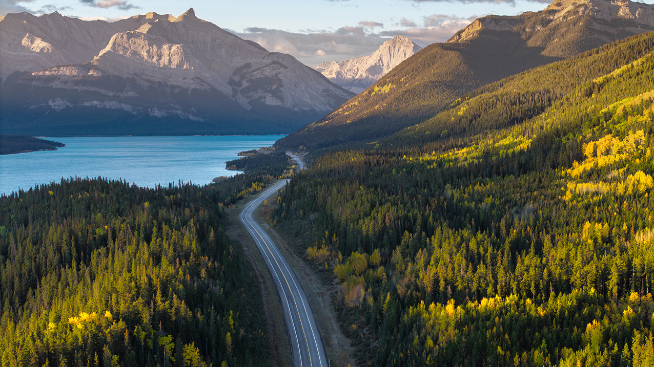Aerial view of a two-lane highway winding through a forest, past a body of water and toward the Rocky mountains. 