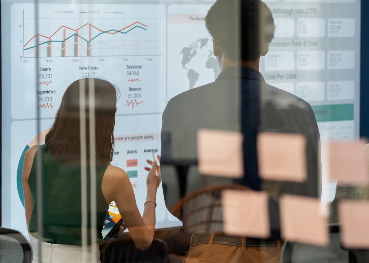 Un groupe de personnes de dos regardant un écran de projection affichant un tableau de données statistiques. On peut voir cette scène à travers une paroi vitrée sur laquelle sont apposés des feuillets autocollants. 